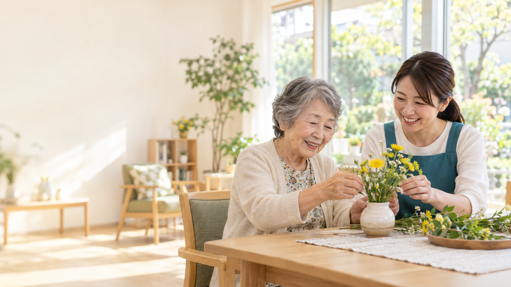 デイサービスの明るい室内で、職員と高齢の女性が季節の花をいける実写調イメージ